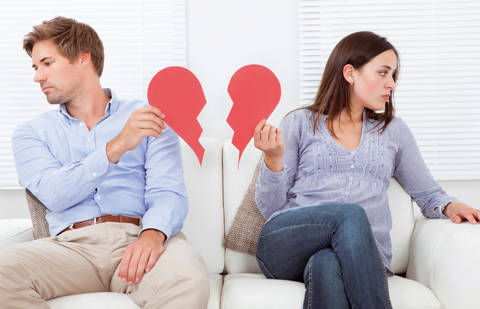 Ein auf der Couch sitzendes Paar das sich voneinander abwendet und jeweils eine Hälfte eines geteiltes roten Herzes in der Hand hält