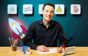 Fabi von Fabis DesignKids zeichnet an einem Schreibtisch und lacht in die Kamera.