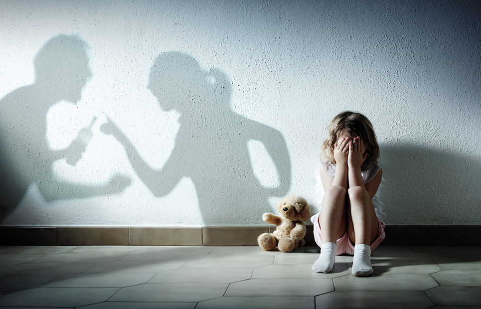 Ein Mädchen sitzt mit den Händen vor Augen auf dem Boden an einer Wand, neben ihr sitzt ihr Teddybär. Man sieht als Schatten auf der Wand die streitenden Eltern. Der Vater hat eine Flasche in der Hand.