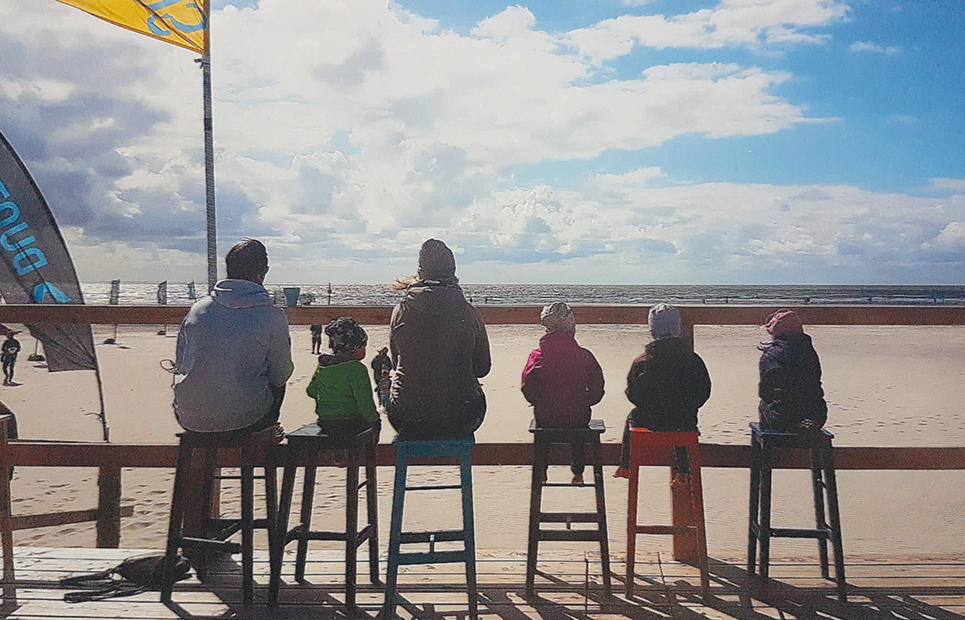 Eine sechsköpfige Familie sitzt mit dem Rücken zur Kamera auf Barhockern auf einer Strandpromenade. Sie schauen aufs Meer hinaus.