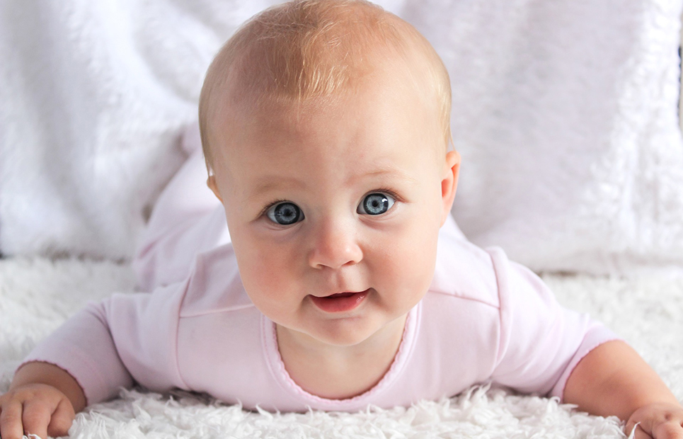 Ein Baby liegt auf dem Bauch, hebt den Kopf und sieht direkt in die Kamera. Es hat strahlend blaue Augen und einen rosafarbenen Strampler an.