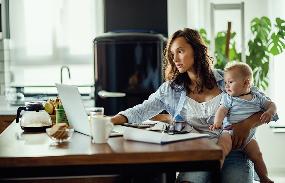 Eine Frau, mit Kleinkind auf dem Arm, sitzt am Schreibtisch und arbeitet am Laptop