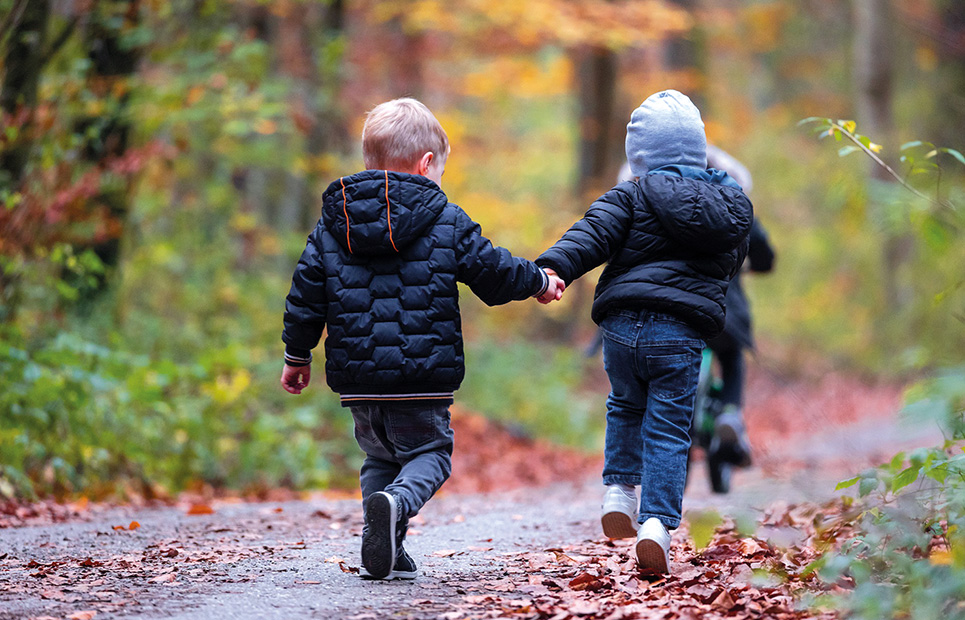 Zwei Kinder im Kindergartenalter laufen Hand in Hand auf einem Weg im Wald